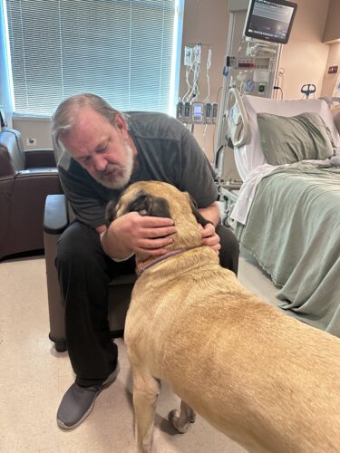 Man comforting dog beside hospital bed.